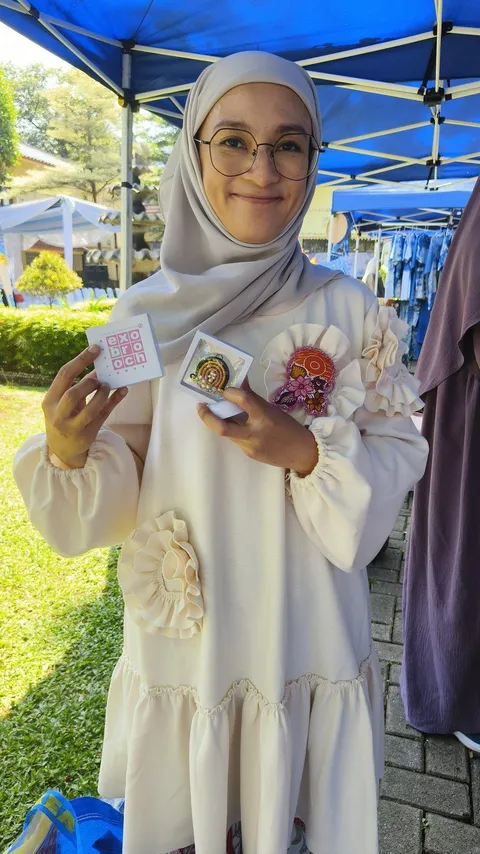 Ika Bunga Maharani tersenyum memegang bros dalam kemasan kartu di bawah tenda pameran