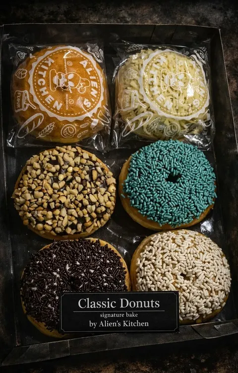 Kotak hitam berisi enam donat bertopping kacang, meses, serta sprinkles biru dan putih, berlabel Classic Donuts
