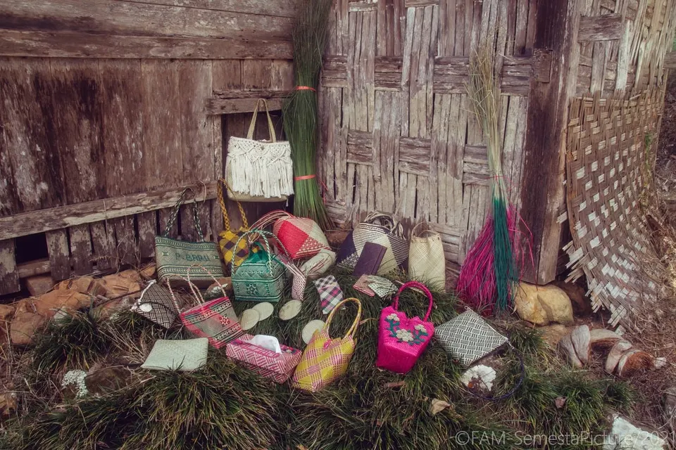 Aneka tas, dompet dan tempat tisu anyaman mansiang warna-warni di atas rumput depan dinding kayu dan bambu