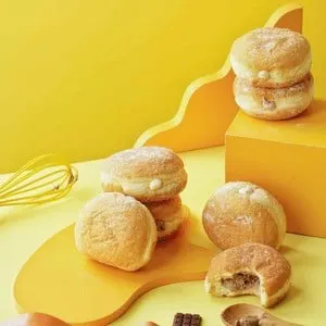 Beberapa donat bertabur gula halus berisi krim di atas latar kuning, salah satunya tergigit berisi cokelat