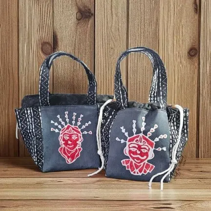 Dua tas jinjing kecil abu-abu bertali serut dengan motif batik ondel-ondel merah di atas meja kayu