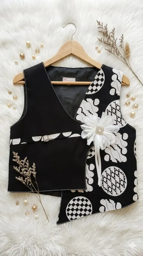 Vest hitam asimetris dengan panel motif batik hitam putih dan bros bunga putih pada gantungan kayu