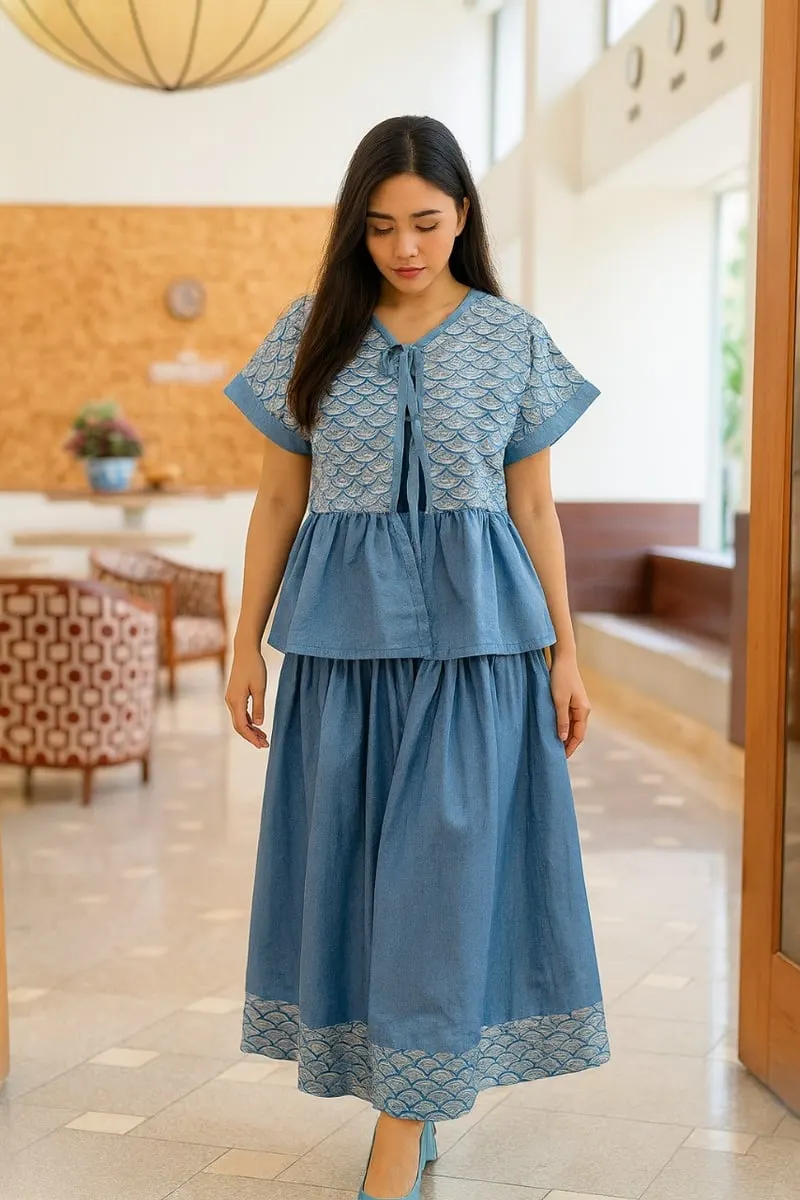 Model perempuan memakai blus biru berpanel motif sisik dan rok biru panjang berlis motif di lobi hotel