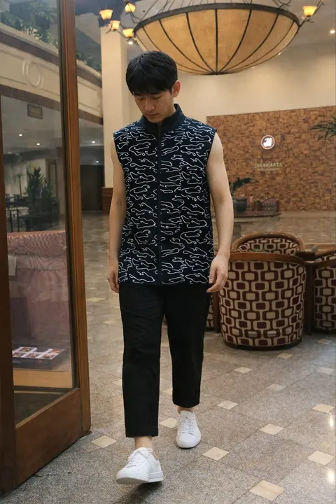 Model pria memakai kemeja tanpa lengan hitam bermotif putih, celana hitam dan sepatu putih di lobi