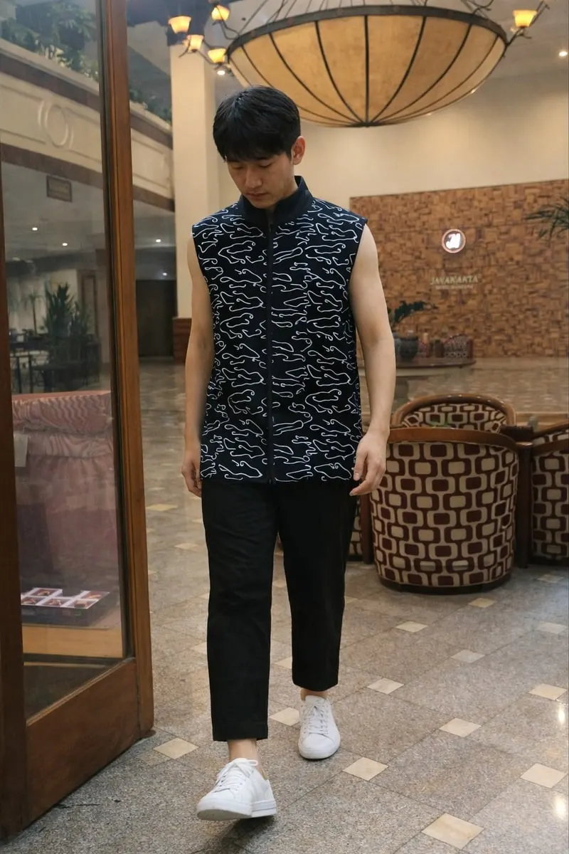 Model pria memakai kemeja tanpa lengan hitam bermotif putih, celana hitam dan sepatu putih di lobi