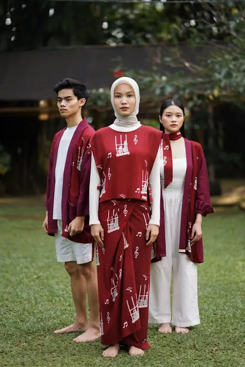 Tiga model di halaman berumput mengenakan busana batik merah marun bermotif angklung