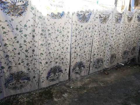 Deretan kain batik putih bermotif sulur daun hijau dan lambang Mosaik dijemur di halaman