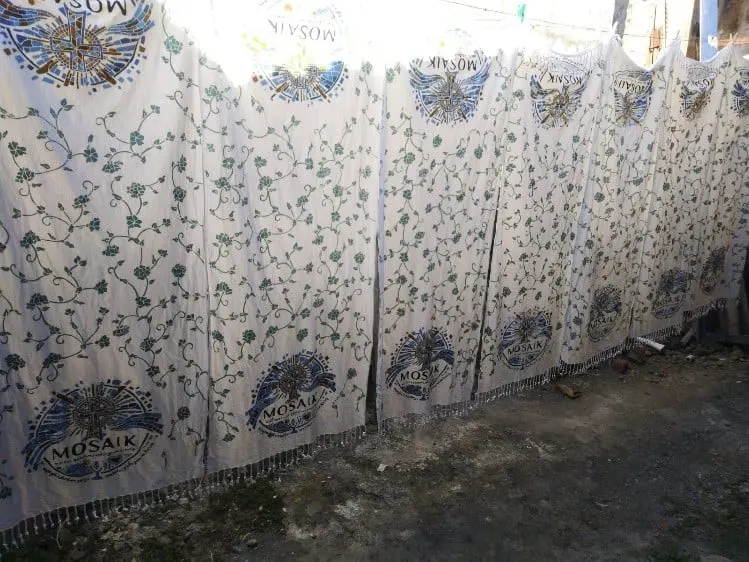 Deretan kain batik putih bermotif sulur daun hijau dan lambang Mosaik dijemur di halaman