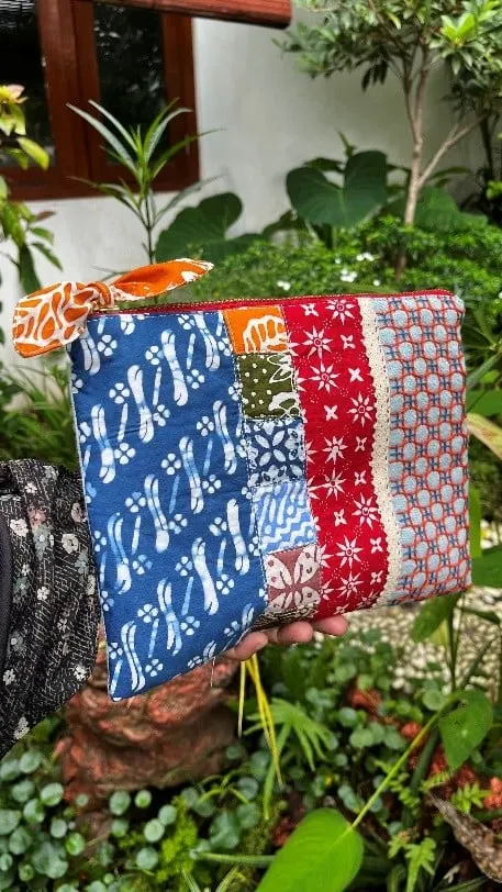 Pouch dari perca batik biru, merah dan putih dengan pita oranye, dipegang di depan tanaman