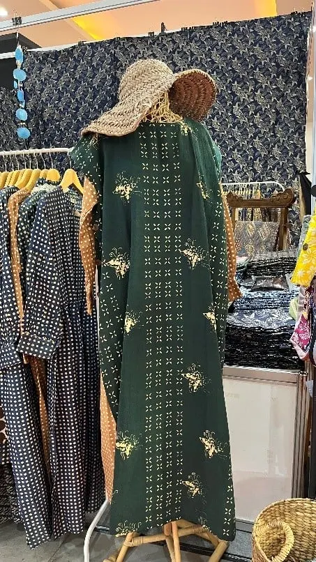 Gaun panjang batik hijau tua di manekin bertopi anyaman, di stan dengan deretan pakaian batik