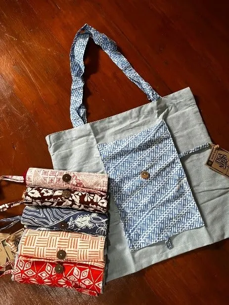Tas belanja kain biru muda dengan saku batik, di samping lima tas lipat batik bergulung berkancing