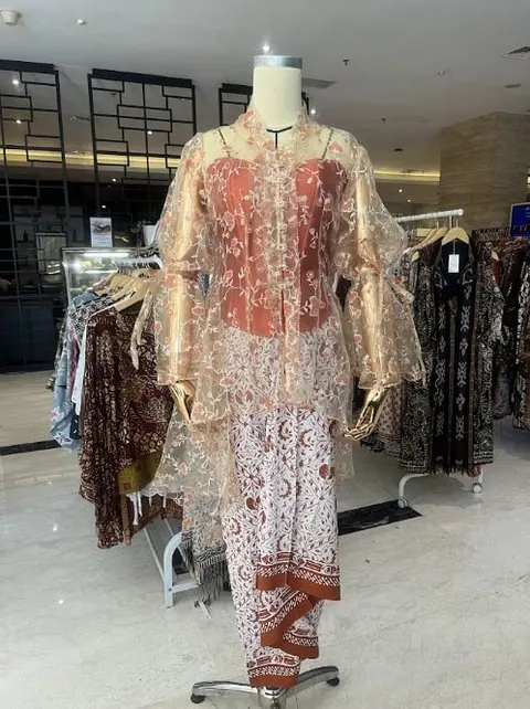 Manekin memakai atasan organza transparan berpotongan kebaya dan celana batik merah bata di butik