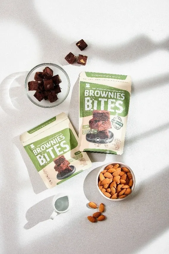 Dua pouch hijau Brownies Bites dengan mangkuk kaca berisi potongan brownies dan mangkuk almon di meja putih