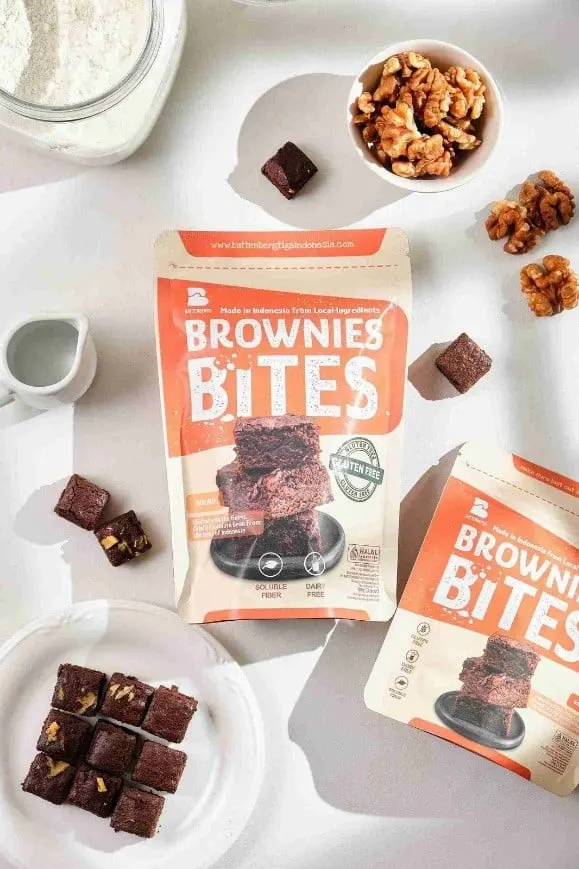 Dua pouch oranye Brownies Bites dengan piring potongan brownies, mangkuk kenari dan teko susu kecil di meja putih
