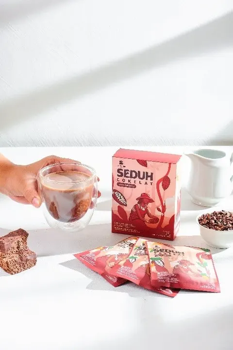 Tangan memegang gelas cokelat di samping kotak merah Seduh Cokelat dan beberapa sachet di meja putih