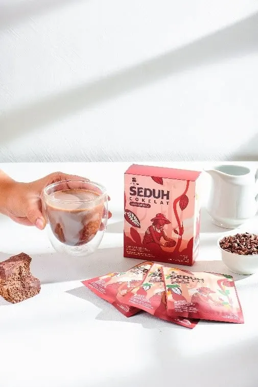 Tangan memegang gelas cokelat di samping kotak merah Seduh Cokelat dan beberapa sachet di meja putih