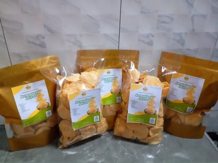 Lima kemasan kemplang beras, dua pouch emas dan tiga plastik bening berlabel hijau kuning, di depan dinding keramik