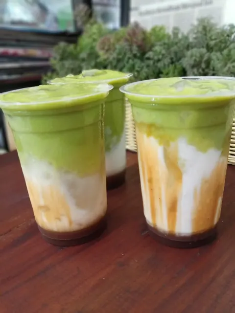 Tiga gelas plastik berisi minuman berlapis hijau, putih dan cokelat di atas meja kayu