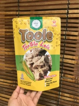 Tangan memegang kemasan pouch kuning Toole Tortila Lele berisi keripik tortila di depan tirai bambu