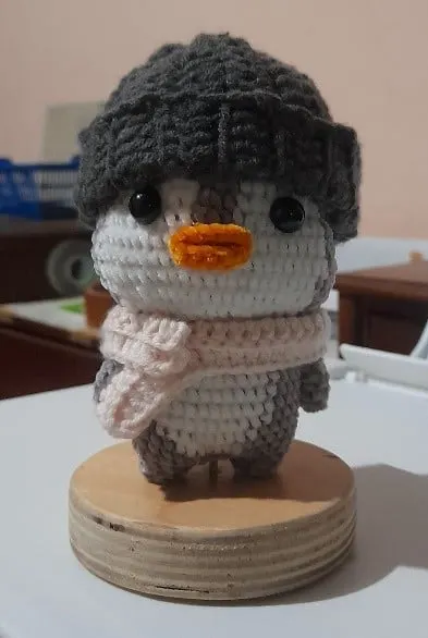 Boneka rajut penguin memakai kupluk abu dan syal merah muda, berdiri di atas alas kayu bundar