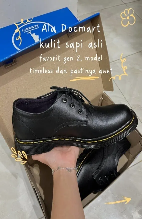 Tangan memegang sepatu kulit hitam bertali dengan jahitan kuning di atas kotak Liberty, dengan tulisan Ala Docmart