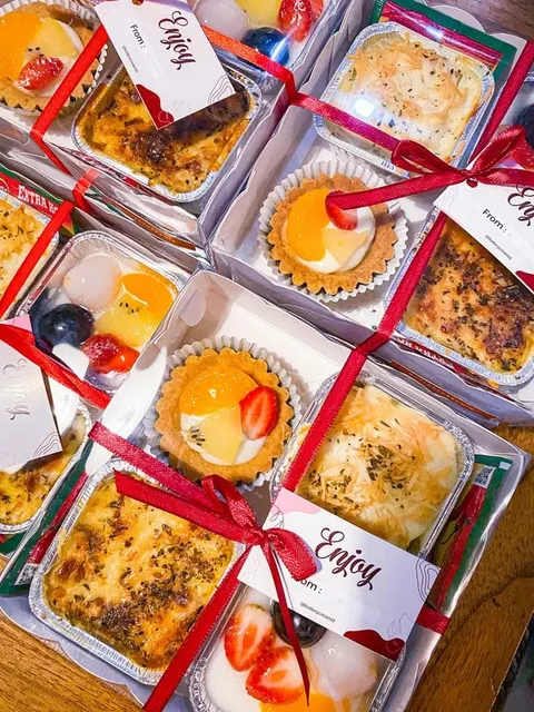 Kotak snack berisi pie buah, hidangan panggang keju dan puding buah, diikat pita merah berlabel Enjoy
