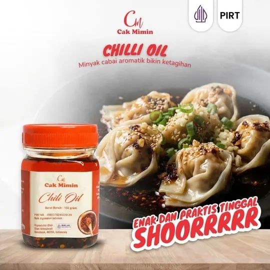 Toples kaca Chili Oil Cak Mimin bertutup merah di depan piring pangsit bertabur minyak cabai