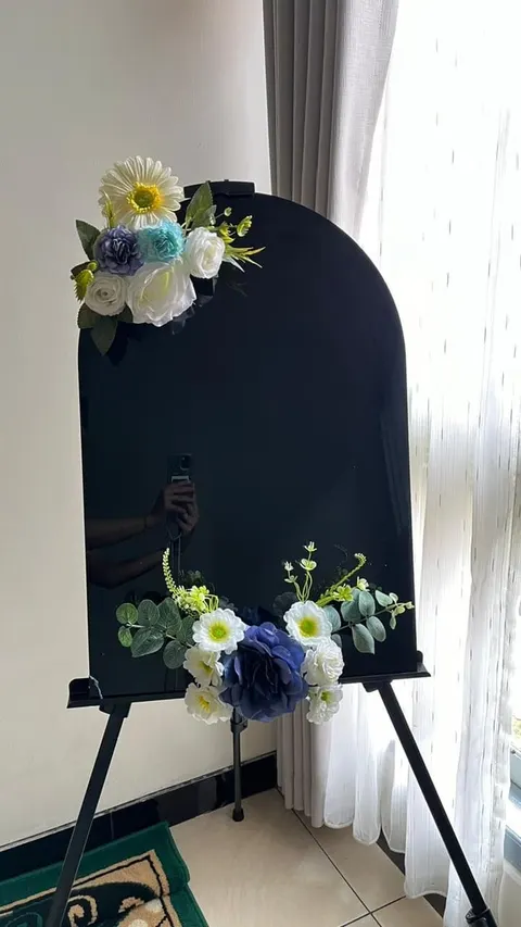 Papan lengkung hitam di atas easel dihiasi rangkaian bunga putih, biru dan daun hijau di dua sudut