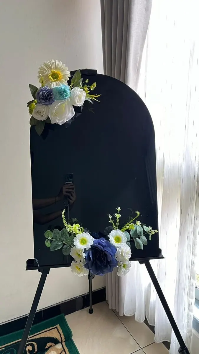 Papan lengkung hitam di atas easel dihiasi rangkaian bunga putih, biru dan daun hijau di dua sudut