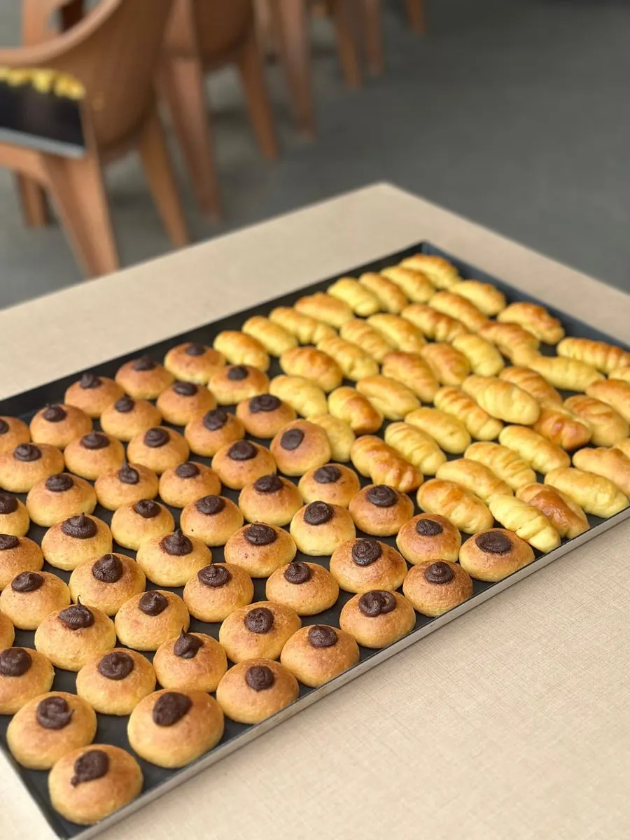 Loyang hitam berisi deretan roti mini bulat bertopping cokelat dan roti gulung kecil keemasan