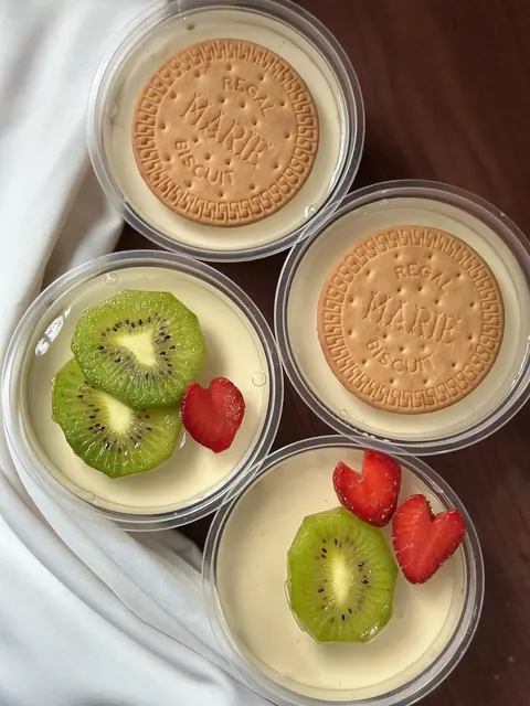 Empat cup plastik berisi dessert krem dengan topping biskuit Marie atau irisan kiwi dan stroberi