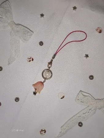 Tali gantungan ponsel merah dengan liontin bunga merah muda dan mutiara di atas kain putih berhias pita renda