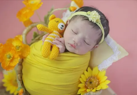Bayi tidur berbalut kain kuning memeluk boneka rajut beruang kuning di antara bunga