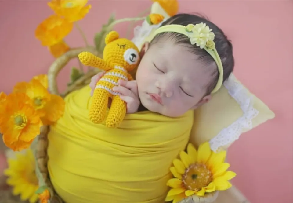 Bayi tidur berbalut kain kuning memeluk boneka rajut beruang kuning di antara bunga
