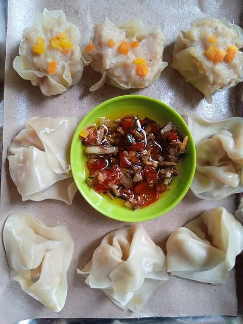 Dimsum bertabur wortel dan pangsit di atas kertas cokelat mengelilingi mangkuk hijau berisi sambal