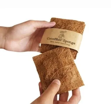 Tangan memegang dua spons sabut kelapa, salah satunya berlabel kertas Cocofiber Sponge