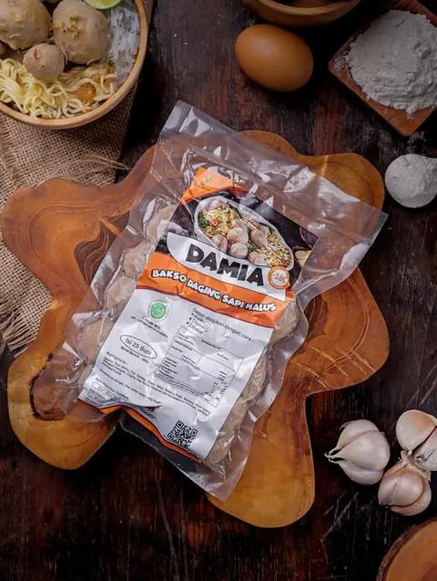 Kemasan vakum Bakso Daging Sapi Halus Damia berlabel oranye di atas talenan kayu, dengan telur dan bawang putih