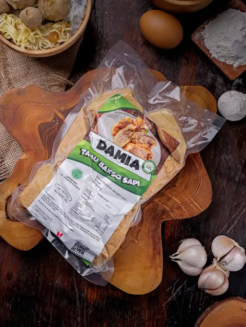 Kemasan vakum Tahu Bakso Sapi Damia berlabel hijau isi 10 pcs di atas talenan kayu, dengan telur dan bawang putih