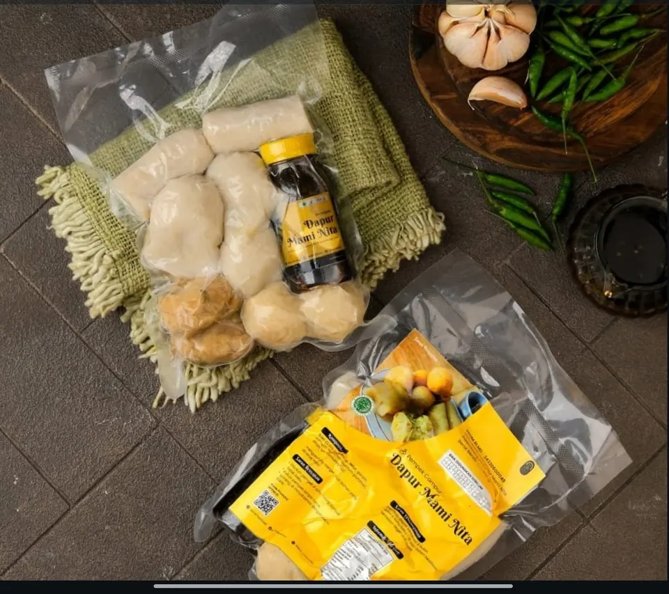 Pempek dalam kemasan vakum bening dengan botol cuko berlabel Dapur Mami Nita dan kemasan kuning di lantai batu