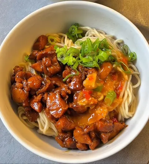 Semangkuk mi dengan potongan ayam bersaus cokelat, saus oranye dan irisan daun bawang