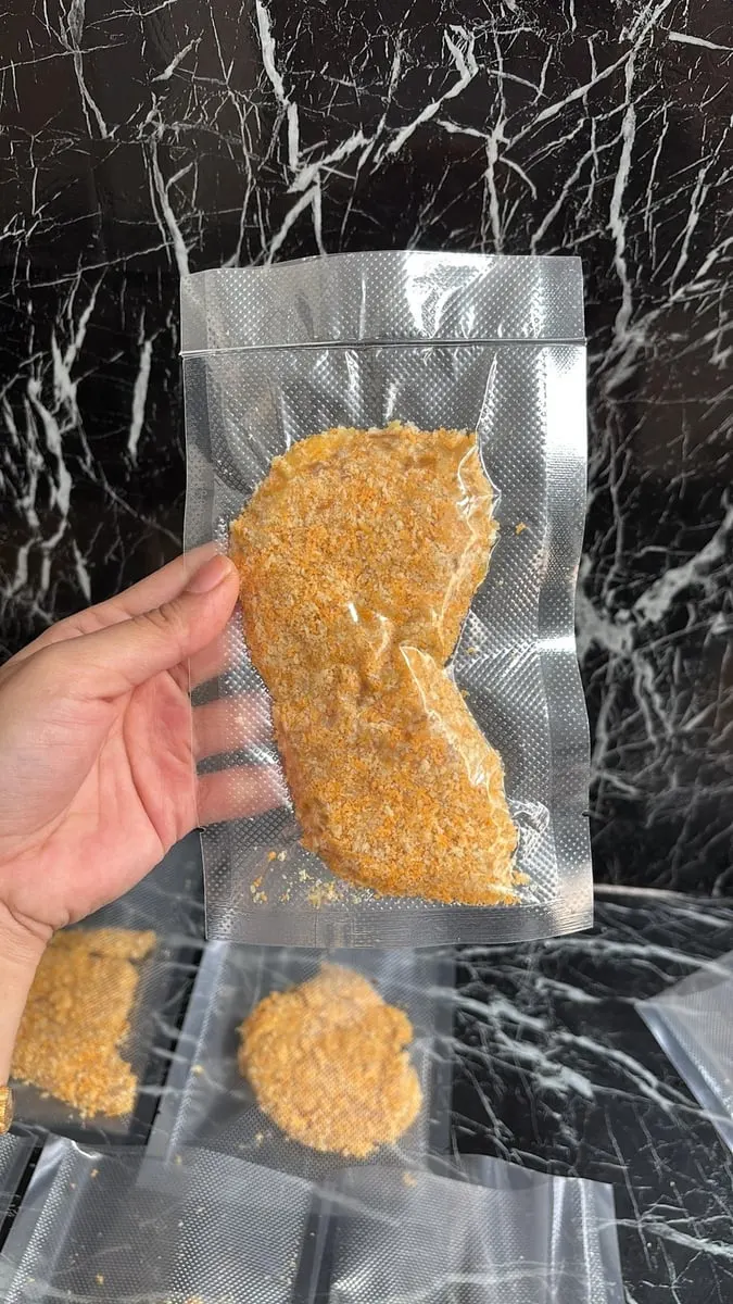 Tangan memegang kemasan plastik vakum berisi lauk berbalut tepung roti di atas meja marmer hitam