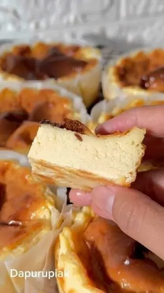 Potongan cheesecake dipegang tangan di atas deretan cheesecake mini berwarna karamel dalam kertas roti