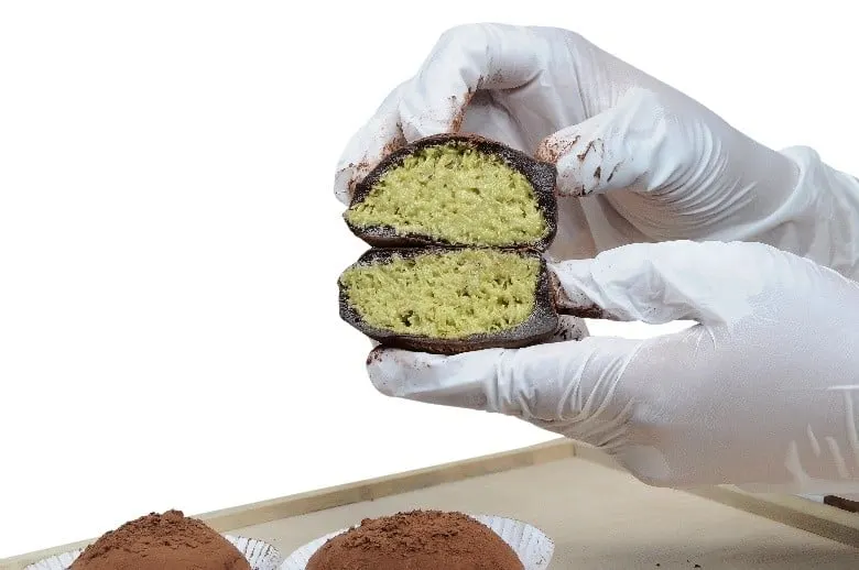 Kue bulat berlapis cokelat dibelah menampakkan isi hijau, dipegang tangan bersarung tangan putih