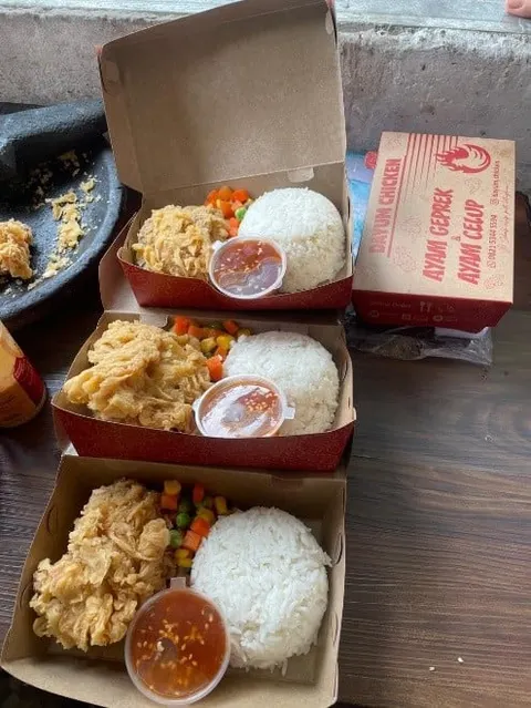 Tiga kotak kertas berisi nasi, ayam goreng tepung, sayur campur dan saus, di samping kotak Dayum Chicken
