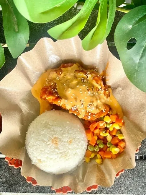Nasi putih, ayam goreng bersiram saus oranye bertabur wijen dan sayur campur di wadah kertas cokelat