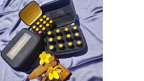 Tas kotak hitam DDistillers berisi botol kecil bertutup kuning di atas kain ungu, dengan bunga kamboja dan kulit kayu