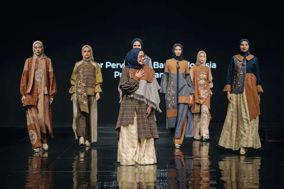 Enam model berhijab dan seorang perempuan berkacamata di runway mengenakan busana tenun cokelat, biru dan krem