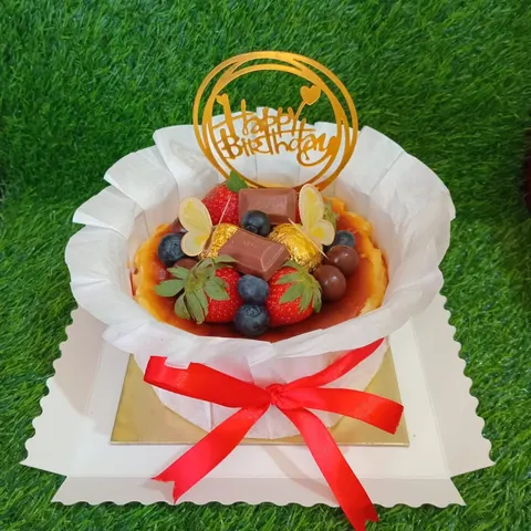 Kue berbalut kertas roti dengan hiasan stroberi, blueberry, cokelat dan topper Happy Birthday, berpita merah