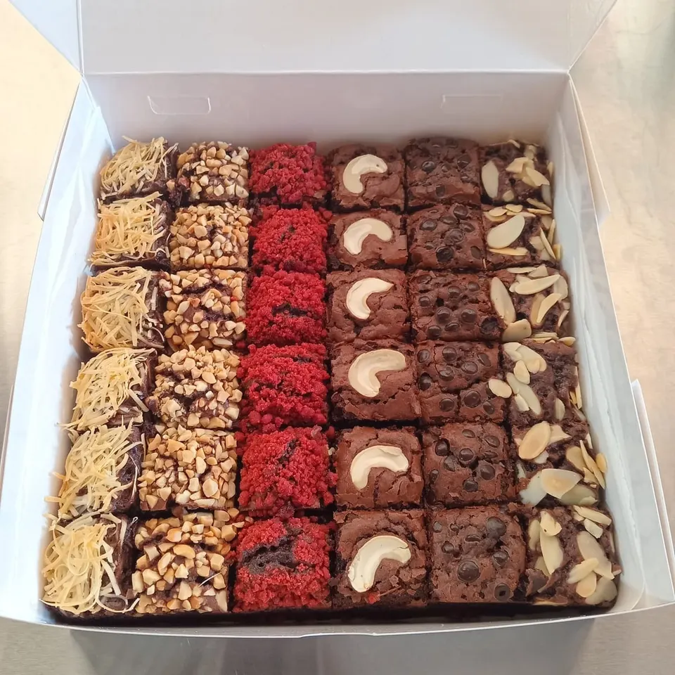 Kotak berisi brownies potong bertopping keju, kacang, remahan merah, kacang mete, choco chip dan almond