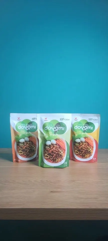 Tiga kemasan pouch Mie Kelor Doyami bergambar mi dan telur di atas meja kayu berlatar dinding biru